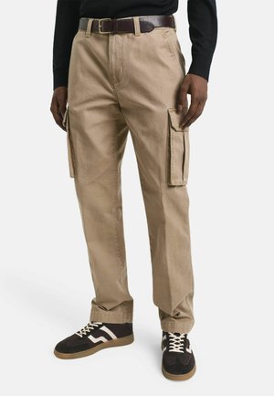 Beige cargobroeken van katoen, met zijzakken, een relaxed fit en een stevige textuur. Draagt met donkere sneakers en een zwart top.