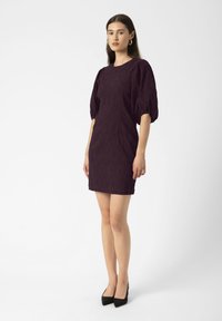 Mujer de pie vistiendo un vestido texturizado de color púrpura oscuro con mangas abullonadas hasta el codo y tacones de puntera negra sobre un fondo blanco liso.