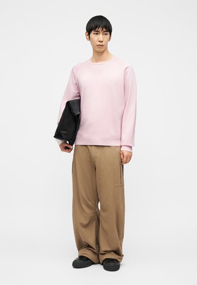 Homme portant un sweat-shirt rose, un pantalon ample couleur beige clair et des chaussures noires, tenant une veste noire pliée sur un fond blanc uni.