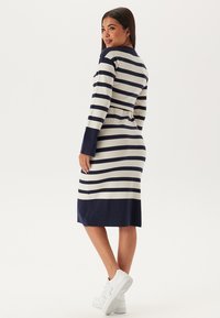 Gestreiftes langärmliges Kleid in Creme und Marineblau. Besitzt eine taillierte Taille, einen knielangen Saum und eine subtile Strickstruktur. Wird mit weißen Sneakers getragen.