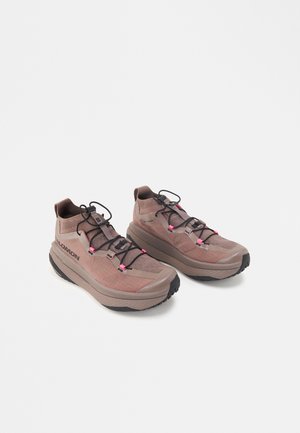 Paar pruune ja roosade Salomoni spordijalatsite paksude tallade ja mustade elastsete paeltega valgel taustal.