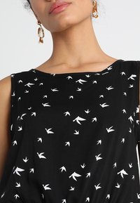 Top nero senza maniche con un motivo di uccelli bianchi sparsi, collo rotondo e texture del tessuto liscia. Accentuato da dettagli di design minimal.