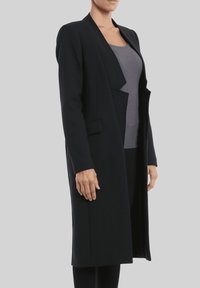 Abrigo largo negro con un frente drapeado, hecho de tela gruesa, con cuello chal y dos bolsillos frontales, usado sobre una blusa gris.