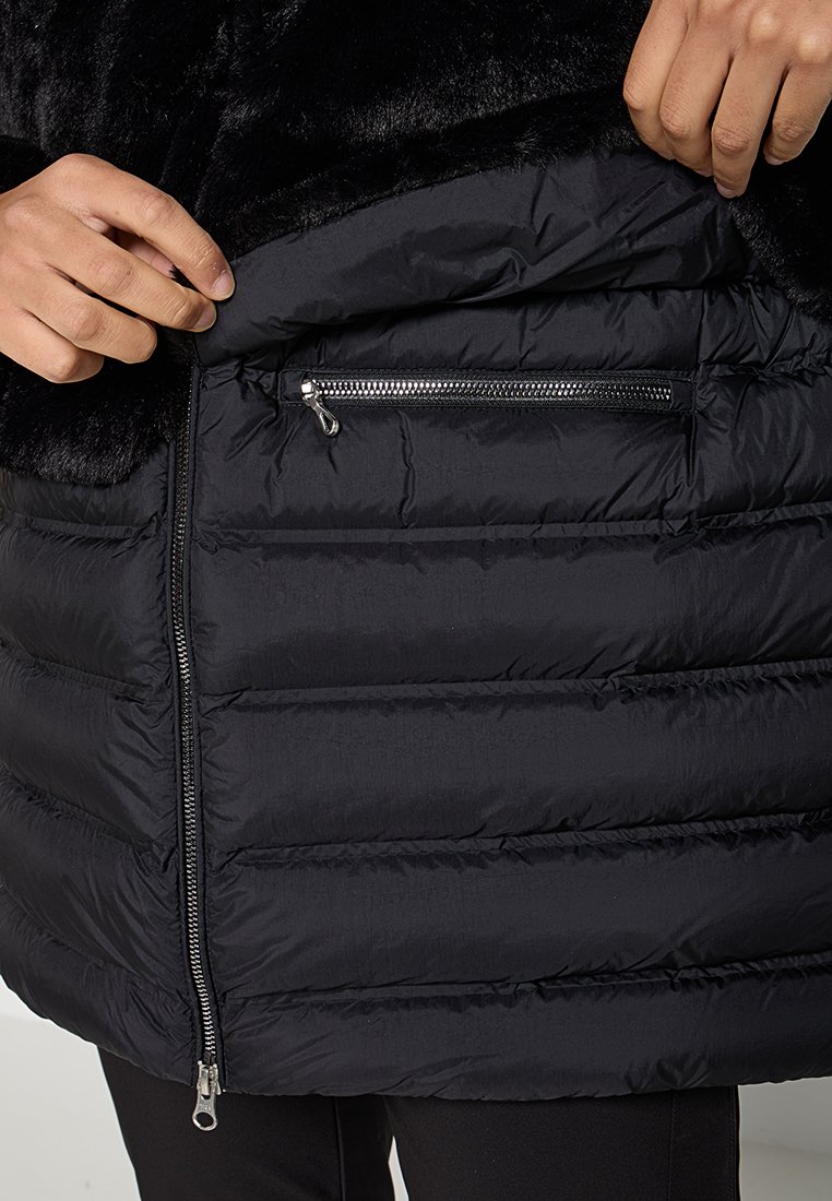 Giacca a vento nera con colletto in pelliccia sintetica, tasca con zip e design trapuntato, finitura lucida e dettagli di cucitura ben visibili.