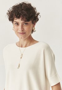 Cremefarvet kortærmet strikket top med løs pasform, parret med guld hoops øreringe og en lagdelt guldvedhængs halskæde. Glat tekstur og minimalistisk design.