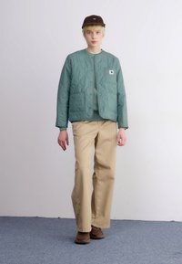 Gequilted groene jas met een ronde halslijn en een vo pocket, gecombineerd met beige broek en bruine schoenen, tegen een neutrale achtergrond.