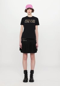 Črna majica s kratkimi rokavi z logotipom "Versace Jeans Couture", v kombinaciji s črno nabranimi krilom in črnimi vojaškimi čevlji; vključuje rožnato kapo.