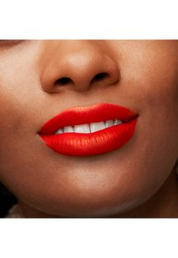 Labios suaves y carnosos con lápiz labial rojo intenso. Dientes blancos y brillantes visibles entre labios cerrados en un tono de piel medio. Superficie texturizada.