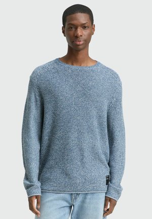 Pull en tricot texturé bleu avec des manches longues, un col rond et une coupe décontractée. Inclus une étiquette sur le côté inférieur. Porté avec un jean bleu clair.