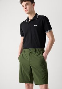 Polo noir avec des accents blancs, arborant un logo, associé à un short vert en tissu léger, doté de poches et d'une taille élastique.
