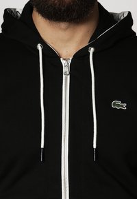 Zwarte zip-up hoodie van zachte stof met een witte rits, voorzien van een groen geborduurd logo en contrasterende witte trekkoorden.