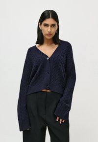 Kvinne iført en mørkeblå kabelstrikket kort cardigan med store knapper og svarte høyt midje bukser, stående mot en enkel lys bakgrunn.