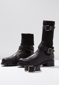Botas de cuero negras hasta el tobillo con una parte superior de tejido acanalado, que cuentan con una correa ajustable y detalles de hebilla metálica. Textura suave y suela gruesa.
