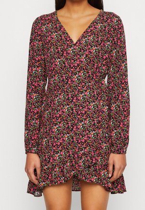 Vrouw draagt een mini-jurk met lange mouwen en V-hals, bedrukt met roze, rood en groene bloemen op een zwarte achtergrond.