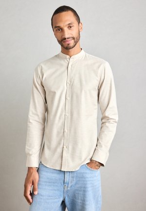 Hombre con cabello corto y barba, vistiendo una camisa de color beige con cuello mandarín y jeans de un tono azul claro, una mano en el bolsillo, fondo neutro.