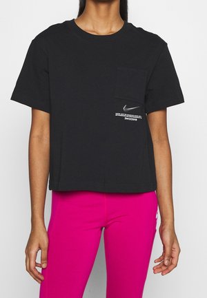 Osoba nosząca czarny krótki t-shirt Nike z przednią kieszenią i małym logo, zestawiony z jaskraworóżowymi legginsami.