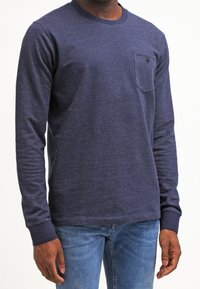 Long-sleeve navy blue knitted shirt with a round neck, front pocket, ribbed cuffs, and a textured pattern. Paired with light blue jeans.