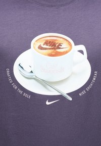 Λευκή κούπα καφέ με την ένδειξη «Café Swoosh» σε πιατάκι, κουτάλι τοποθετημένο στο πιατάκι, τέχνη λατέ που εμφανίζει το «NIKE» και το λογότυπο swoosh, κείμενο που αναγράφει «Κατασκευασμένο για την σόλα.»