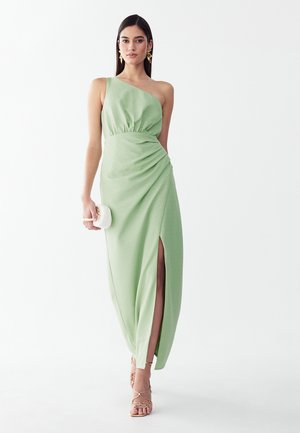 Frau trägt ein hellgrünes One-Shoulder-Kleid mit einem hohen Schlitz, hält eine kleine weiße Tasche und trägt goldene Riemchenabsätze und Ohrringe.