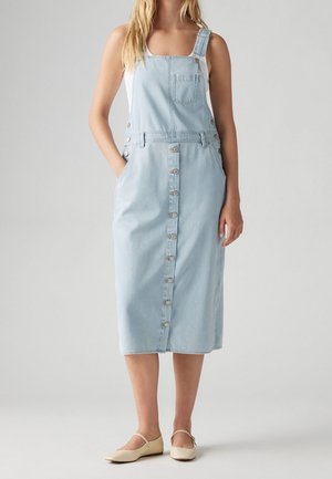 Woman wearing a light blue denim button-front overall dress with pockets and beige flat shoes, standing against a plain background.