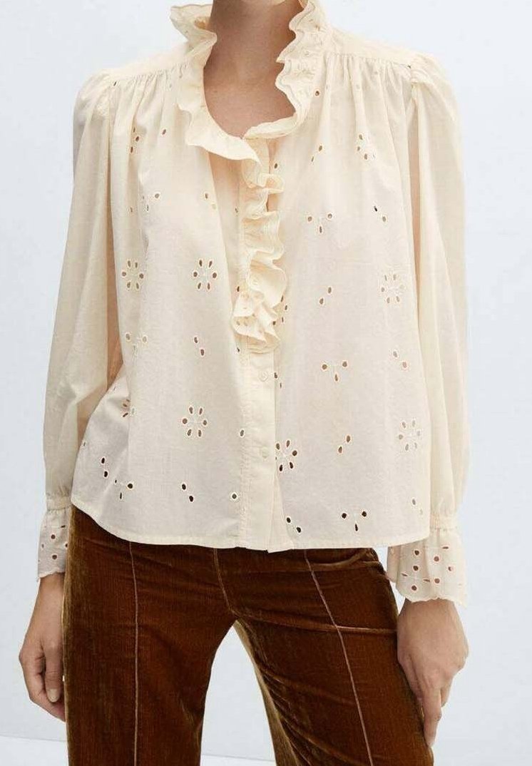 Blusa di cotone color crema con scollo ruffled e chiusura con bottoni, caratterizzata da motivi floreali intagliati e maniche lunghe a sbuffo.