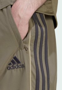Olivovo zelené športové šortky s otvorom na zips a tromi čiernymi vertikálnymi pruhmi, s čiernym logom Adidas vpredu.