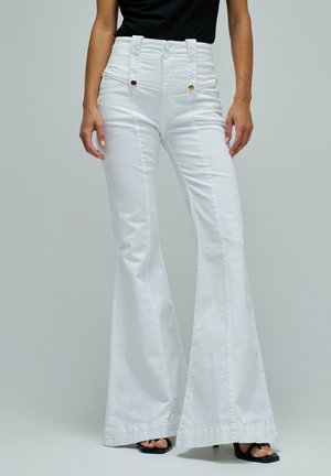 Femme portant un pantalon flare blanc taille haute avec bretelles boutonnées et des talons ouverts noirs, debout devant un fond gris uni.