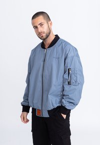 Veste bomber bleu clair avec une fermeture éclair diagonale, des poignets côtelés noirs et un petit logo violet sur la poitrine. Elle possède une poche sur le bras avec fermeture éclair.