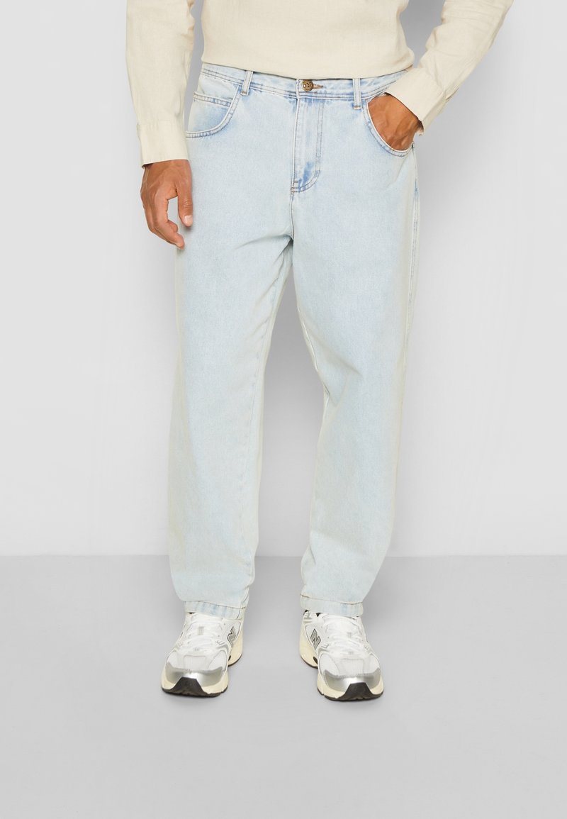 Jean en denim bleu clair avec une coupe ample, design à cinq poches et détails de couture visibles. Porté avec des baskets blanches.
