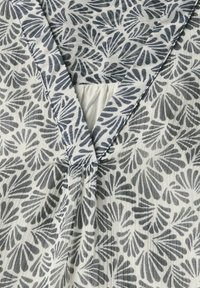 Valkoinen kangas, jossa on tummansininen lehtikuviotulostus. Ominaisuuksiin kuuluu V-mallinen kaula-aukko ja poimutus etuosassa, pehmeä ja teksturoitu pinta.