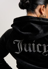 Crna baršunasta dukserica s kamenčićima koji oblikuju riječ "Juicy" na leđima. Kapuljača udobno sjedi na ovratniku.