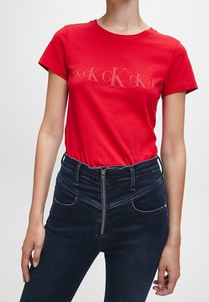 Weiblicher Torso trägt ein rotes T-Shirt mit kurzen Ärmeln und "CK"-Logo auf der Brust, kombiniert mit hoch taillierten dunkelblauen Jeans mit vorderem Reißverschluss.