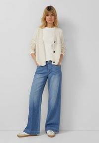 Cardigan in cremiger Textur mit schwarzen Knöpfen, getragen über einem weißen, gerippten Oberteil, kombiniert mit weit geschnittenen hellblauen Jeans und weißen Turnschuhen.