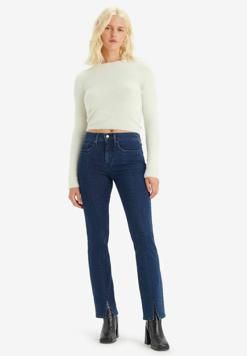 Maglione cropped bianco e peloso abbinato a jeans a vita alta blu scuro con fessure laterali, indossato con stivali neri alla caviglia.