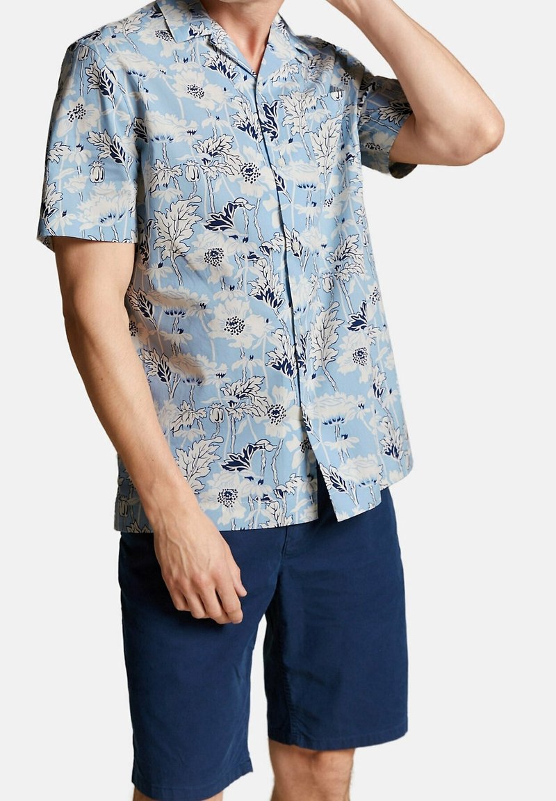 Homme portant une chemise à manches courtes à boutons bleu clair à motif floral et un short bleu foncé, debout avec une main près de son cou.
