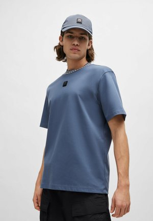 Blue cotton t-shirt with a round neckline and a textured gray collar, featuring a small black logo on the front. Worn with a matching cap.