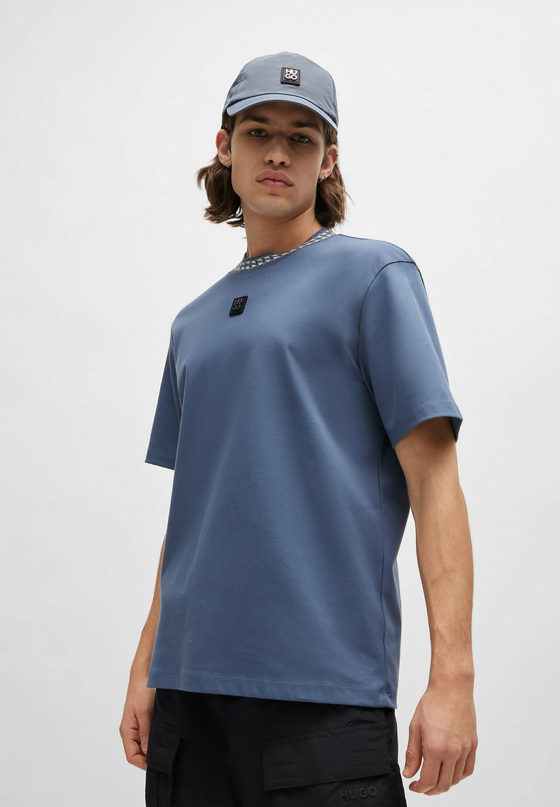 Camiseta de algodón azul con cuello redondo y un cuello texturizado gris, con un pequeño logo negro en la parte delantera. Llevada con una gorra a juego.