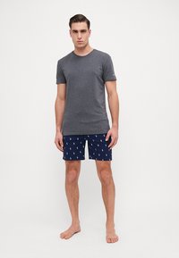 T-shirt gris à manches courtes associé à un short de bain bleu marine avec des imprimés de joueur de polo blancs. Le mannequin se tient pieds nus, les mains sur les hanches.
