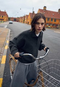 Mörkgrå oversized tröja i ullblandning med krage, tillsammans med ljusgrå, veckade mjukisbyxor. Personen håller en cykel med en trådkorg.