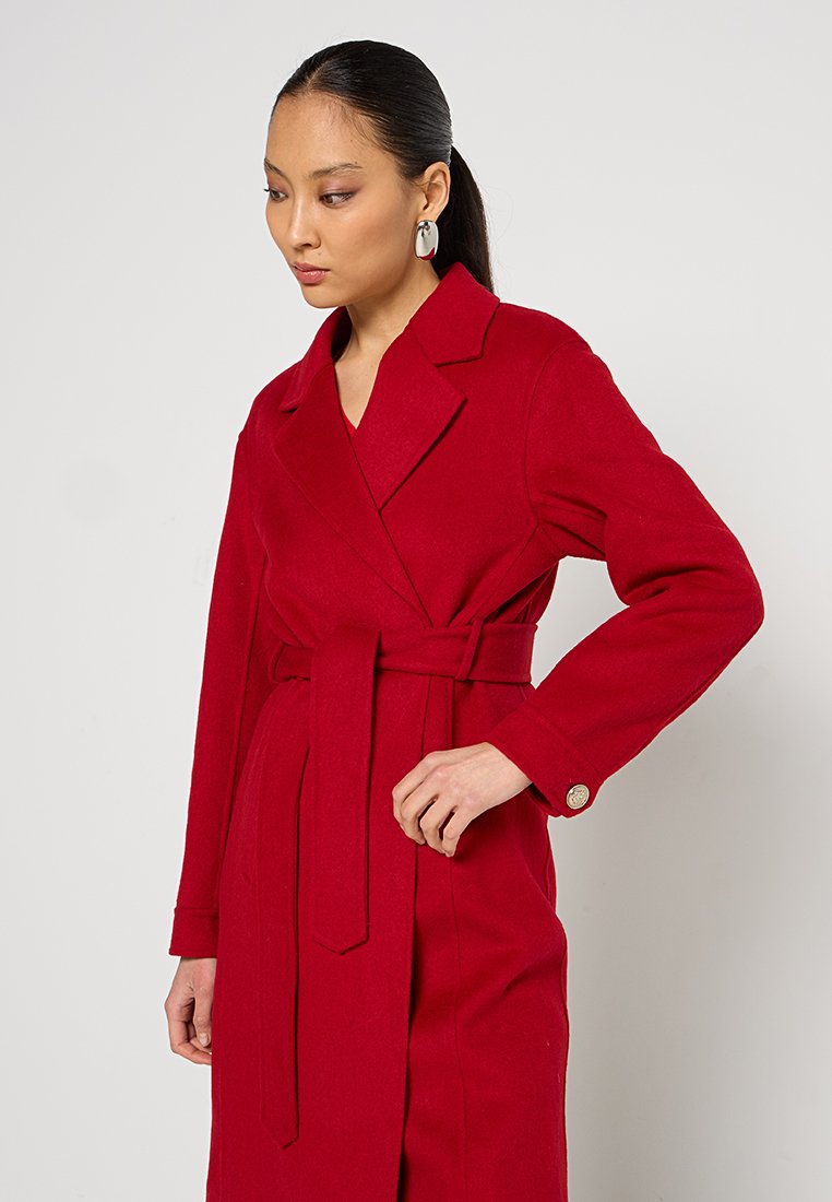 Roter Woll-Trenchcoat mit Gürtel, Reverskragen und geknöpften Manschetten. Er zeichnet sich durch ein elegantes Design und sichtbare Textur aus.