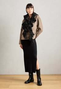 Brun uldblandingsjakke med rynket sort tørklæde; sort ribbet midiskirt med sideskille; sorte læderankelstøvler; trægulv som baggrund.