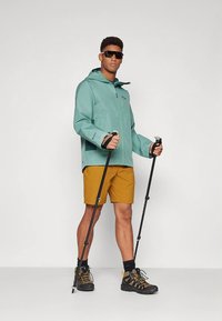 Chaqueta impermeable color teal con capucha, combinada con pantalones cortos mostaza. El modelo sostiene bastones de trekking, lleva gafas de sol oscuras y botas de montaña.