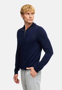 Pullover a maglia navy con colletto a zip, maniche lunghe e una vestibilità rilassata; abbinato a pantaloni tuta grigio chiaro. Tessuto liscio.