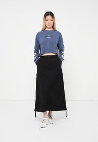 Kratka modra trenirka z dolgimi rokavi, ki vsebuje črno grafiko; kombinirana z dolgo črno cargo krilo in belim obutvijo.