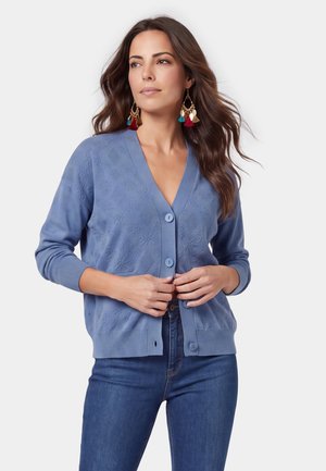 Vrouw met lang bruin haar, die een blauw patroon vest met knopen draagt, blauwe jeans en kleurrijke kwastjesoorbellen, kijkt opzij.