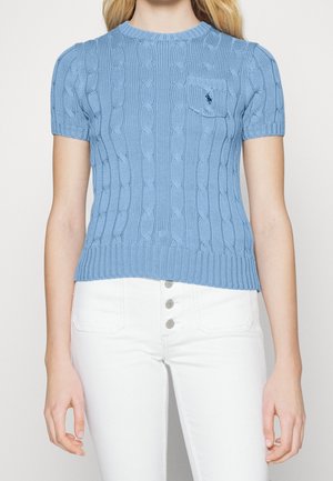 Vrouw draagt een lichtblauwe kabelgebreide trui met korte mouwen, een klein borstzakje en witte broek met knopen.