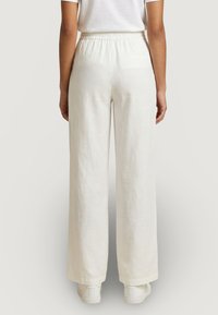 Personne portant un pantalon blanc ample avec une taille élastique, associé à des baskets blanches, vue de dos sur un fond clair.