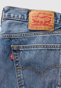 Denim jeans med en lys blå vask, der har en lædermærkeetiket, orange syninger og et lille rødt mærke på baglommen.