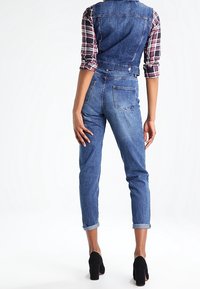 Denim overall met verstelbare bandjes, medium blauwe wassing, taps toelopende pijpen, omgeslagen mouwen en achterzakken. Draagt over een geruite blouse.