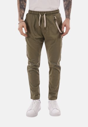 Uomo con pantaloni verde oliva con coulisse e tasca laterale con cerniera, indossa scarpe da ginnastica bianche, mani rilassate lungo i fianchi, tatuaggi visibili sugli avambracci.